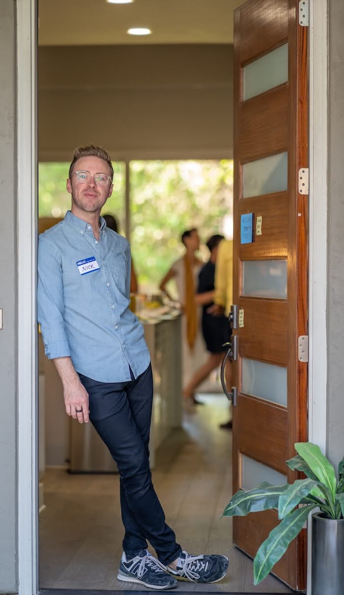 Nick Gray in a doorway wearing a HELLO NICK name tag, a party behind him